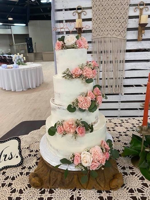 Three-tier white wedding cake with pink roses and greenery on a rustic wooden base.