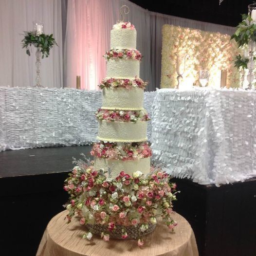 Tall white wedding cake with pink floral decorations on a table, in a decorated reception hall