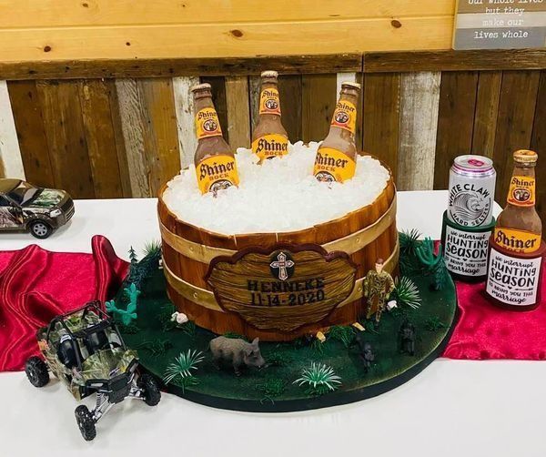 Beer-themed cake with bottles and cans on a green base, displayed on a table with toy tractors and a red cloth.