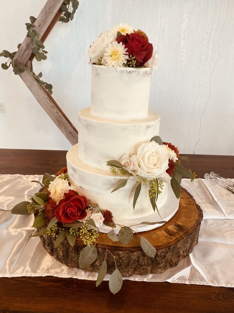 Three-tier white wedding cake with red and white flowers on a rustic wood stand