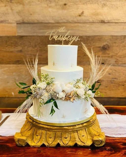 Two-tier white wedding cake with florals on a gold stand against a rustic wooden backdrop