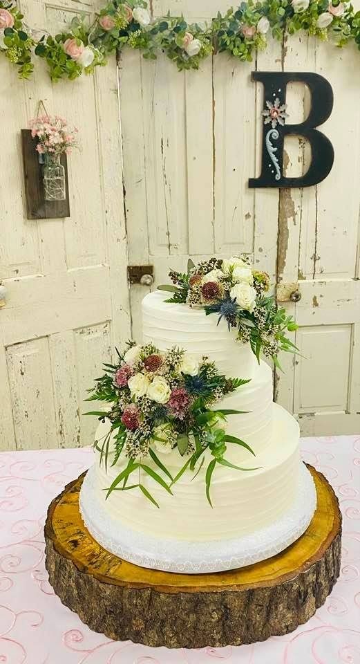 Three-tier white wedding cake with floral greenery, displayed on a tree-slice stand against a rustic backdrop