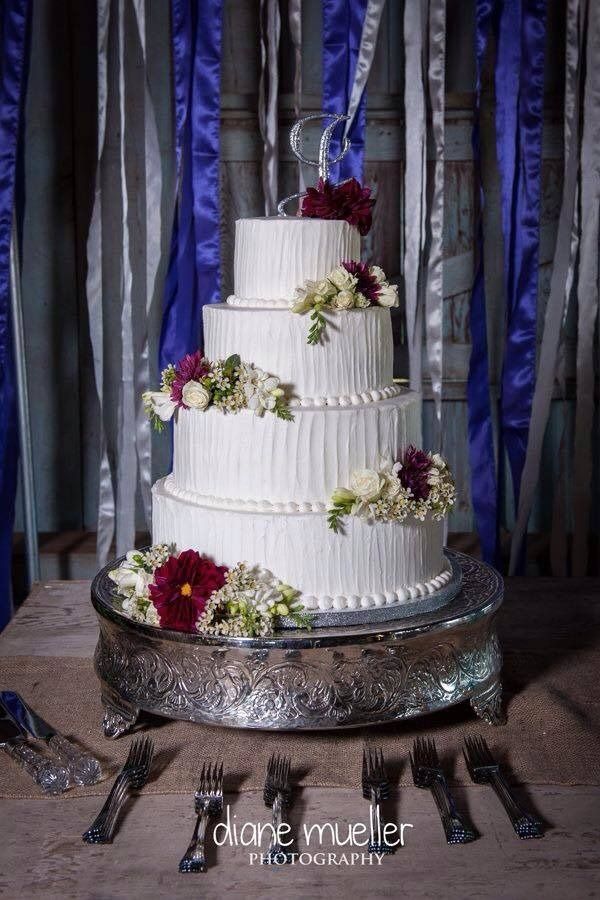 Four-tier white wedding cake with purple and white flowers on a silver stand, set on a decorated table.