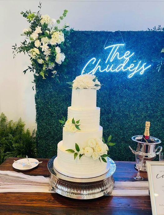 Three-tier white wedding cake with flowers on a table beside a neon “The Chandeys” sign