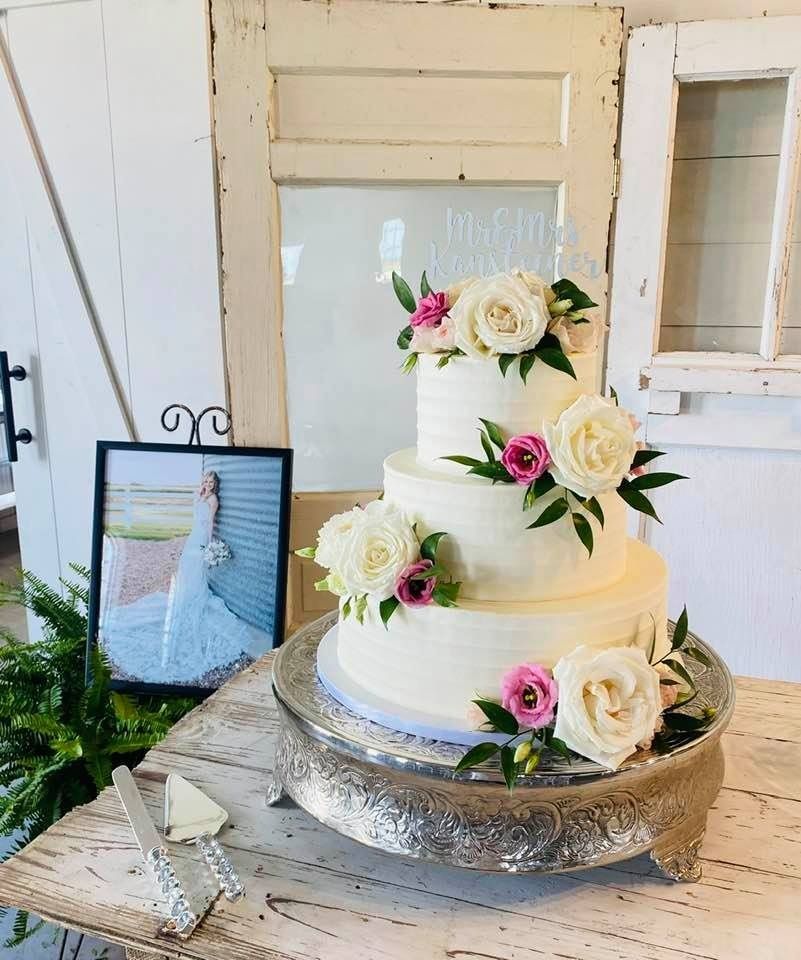 Four-tier white wedding cake with white and pink flowers on a silver stand beside a framed bride photo