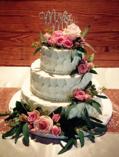 Three-tier white wedding cake with pink roses and greenery on a wooden backdrop