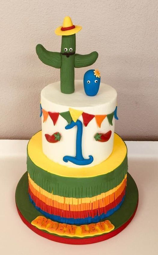 Three-tier cactus-themed birthday cake with rainbow icing, blue number 1, and small decorations
