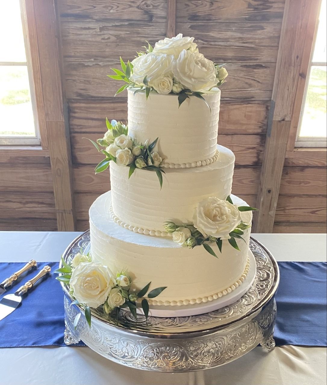 Three-tier white wedding cake with floral decorations on a silver stand in a rustic wood setting
