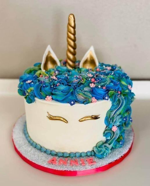 Unicorn cake with blue icing, gold horn and ears, and red letters on a white stand