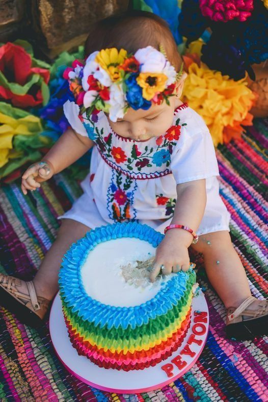Child in white embroidered outfit with flower crown, seated beside colorful woven mat and basket of white petals