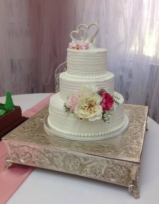 Three-tier white wedding cake with floral decorations and heart topper on a silver stand
