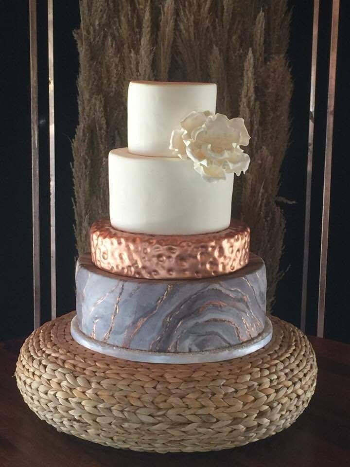 Two-tier white cake with floral accent on copper and gray stands over a woven round base