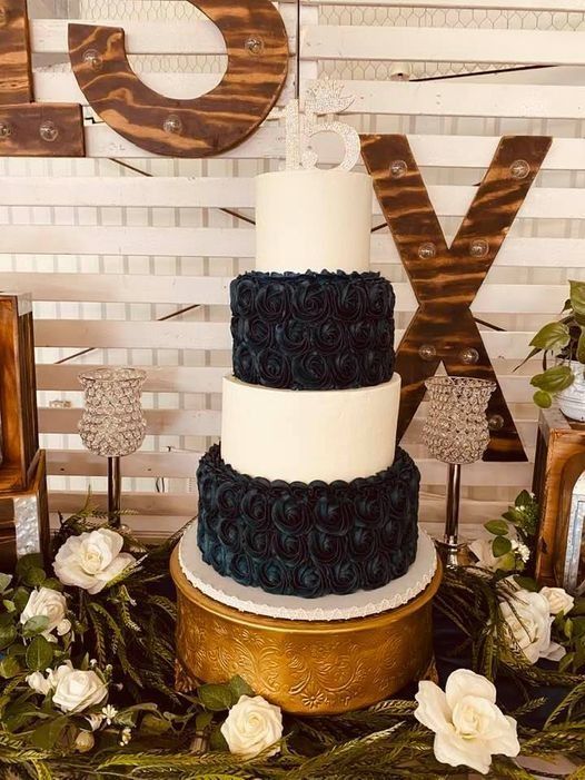Three-tier white and black wedding cake with gold base and floral decorations on a rustic backdrop