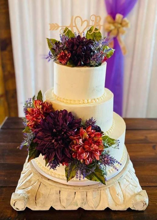 Three-tier white wedding cake with red and purple floral decorations on a carved table centerpiece
