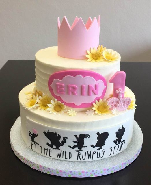 Two-tier white birthday cake with pink crown topper, “ERIN” plaque, and “LET THE WILD RUMPUS START” text