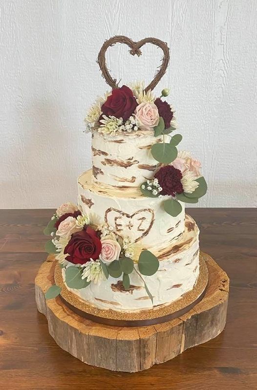 Three-tier rustic wedding cake with white frosting, burgundy and cream flowers, and a heart topper on a wooden slab