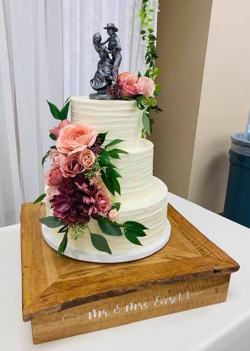Three-tier white wedding cake with pink flowers and a silver topper on a wooden stand