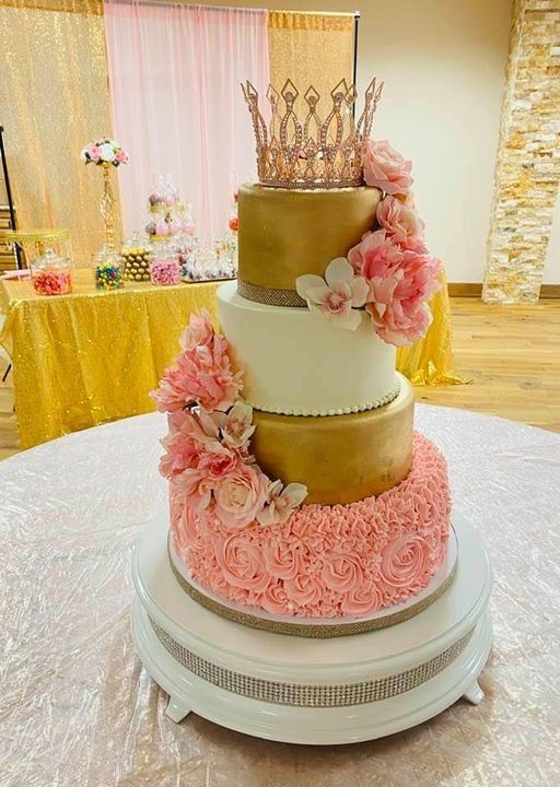 Tall gold, white, and pink tiered wedding cake with a crown topper and floral decorations at a reception table