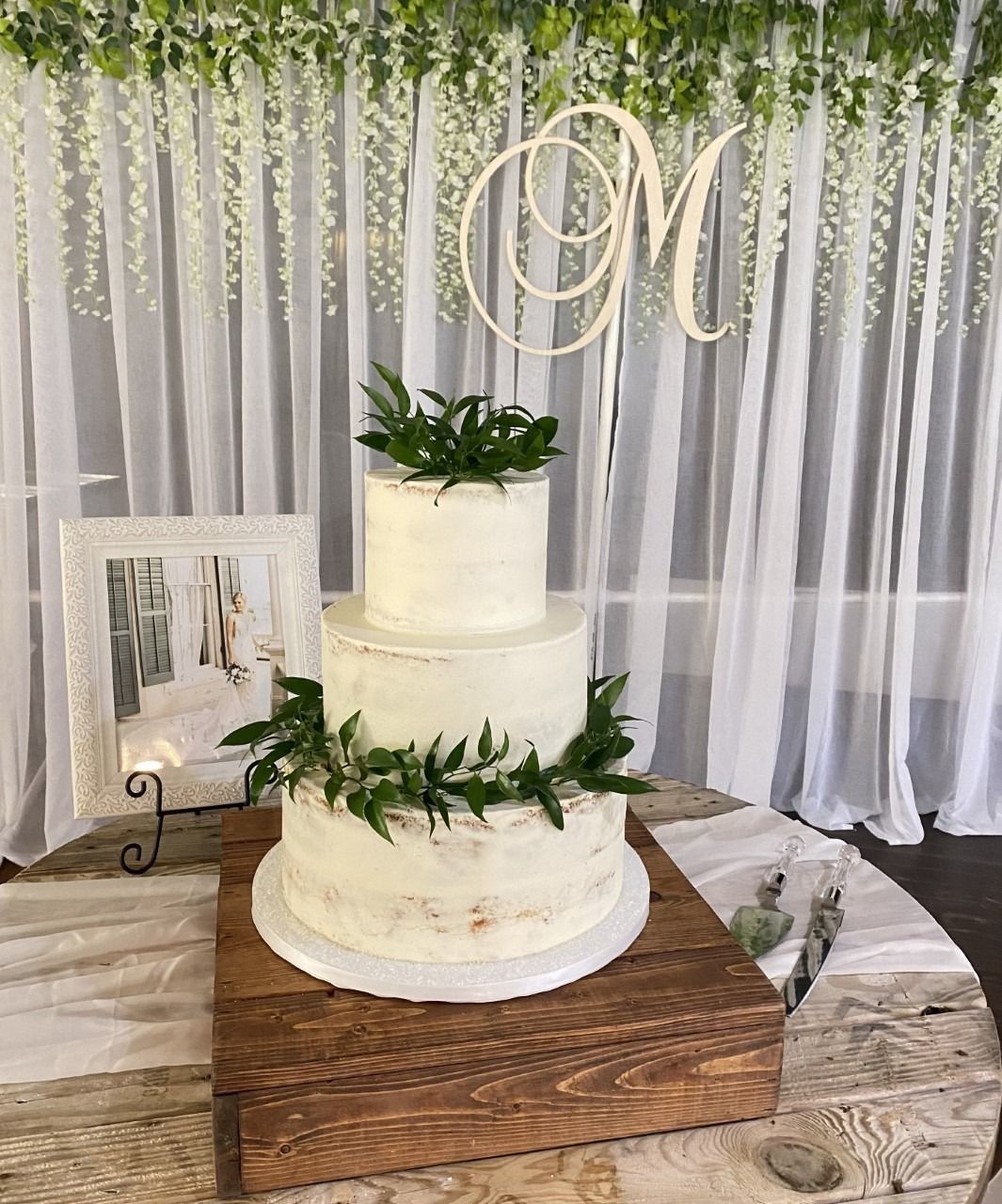 Three-tier white wedding cake with greenery on a wooden stand before a sheer draped backdrop