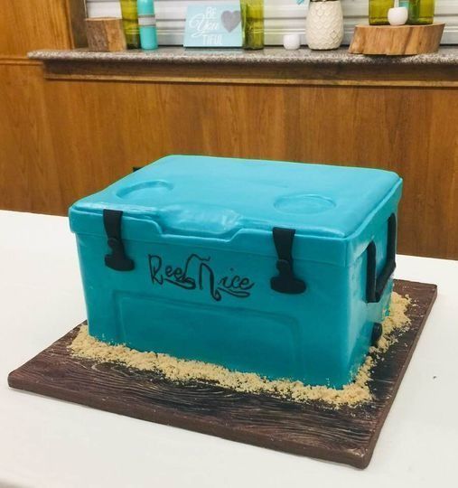 Turquoise suitcase-shaped cake on a wooden board, with black straps and writing on top.