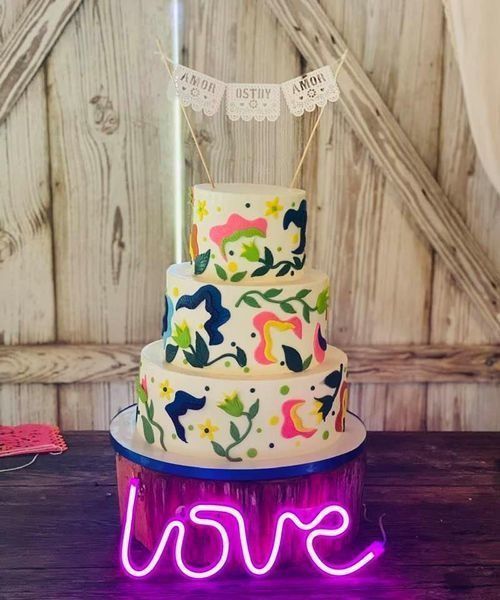 Three-tier floral cake with neon pink “love” sign in front, on a rustic wooden backdrop