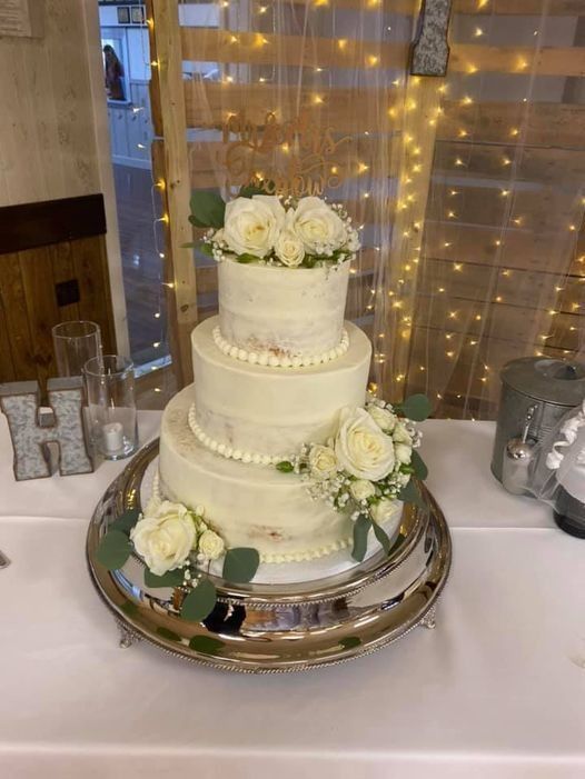 Three-tier white wedding cake with white roses on a silver tray, lit by warm string lights