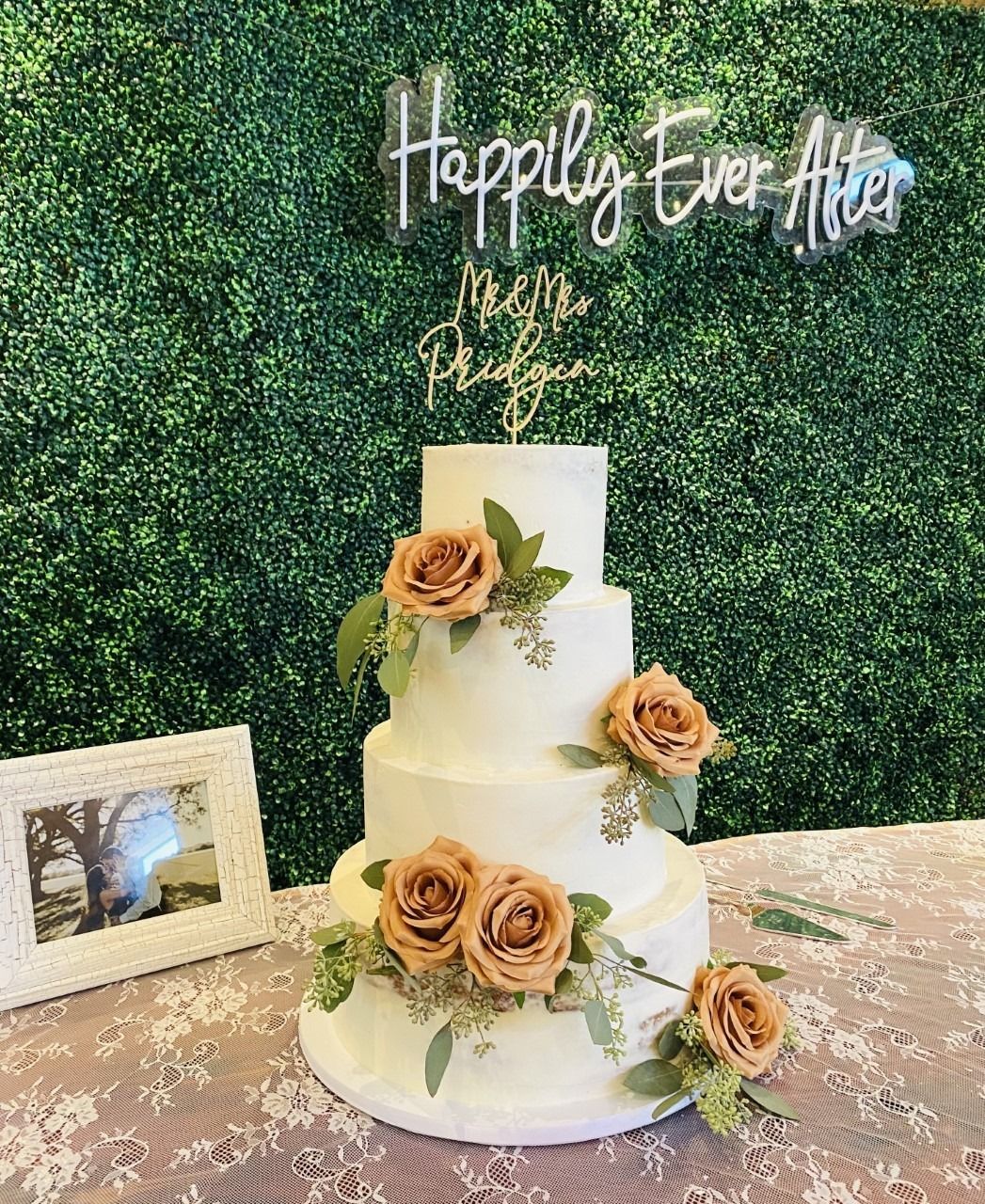 Two-tier white wedding cake with peach roses and greenery under a “Happy Ever After” sign