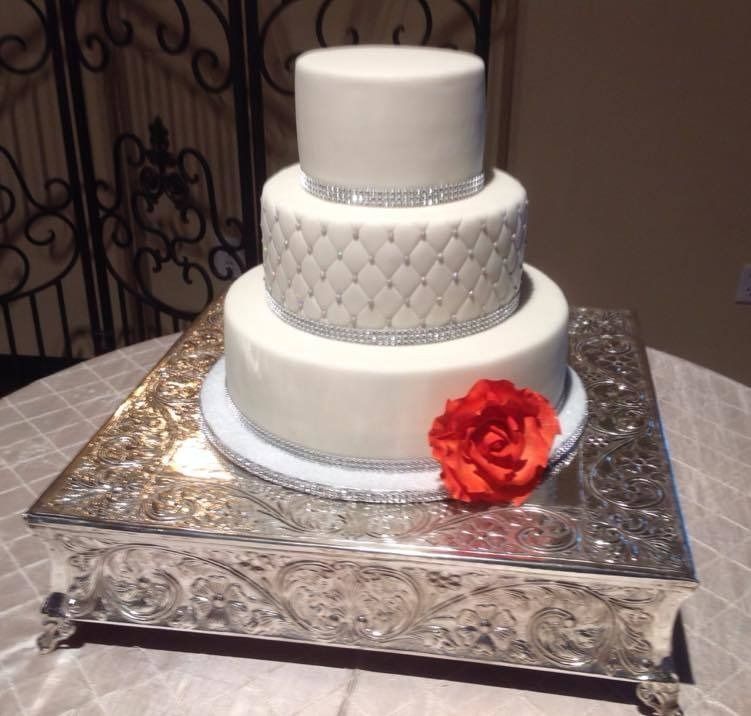 Three-tier white wedding cake with silver accents and a red flower on a silver square stand