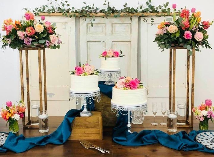 Floral wedding dessert display with white cakes on wooden stands and pink, peach, and blue flowers
