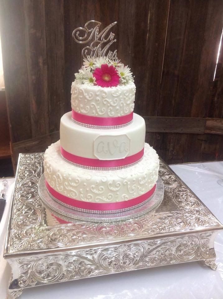 Three-tier white cake with pink trim and a flower topper on an ornate silver stand
