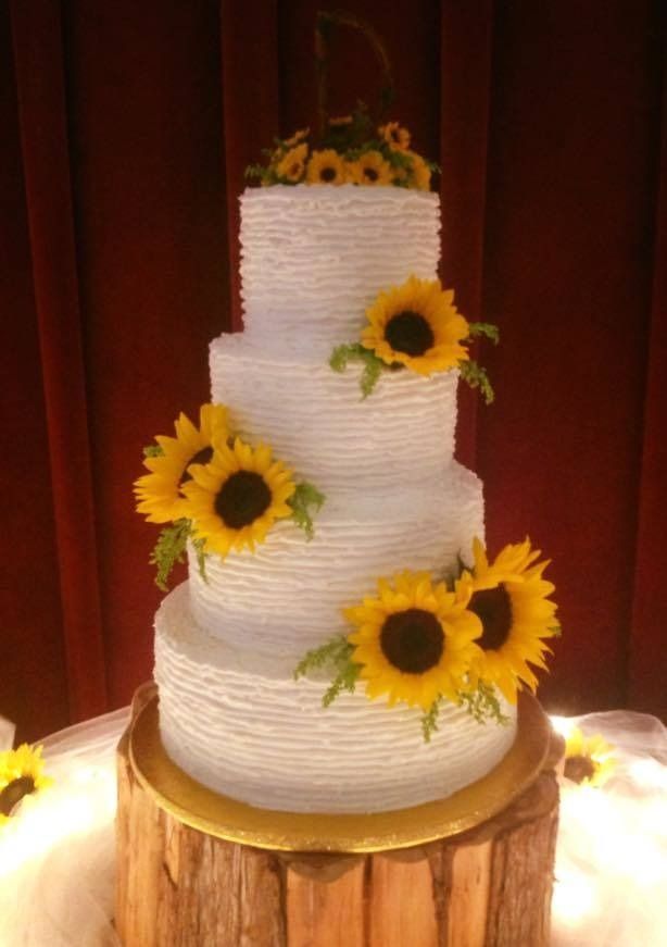 White tiered cake with yellow sunflowers on a wooden stand against a red curtain backdrop