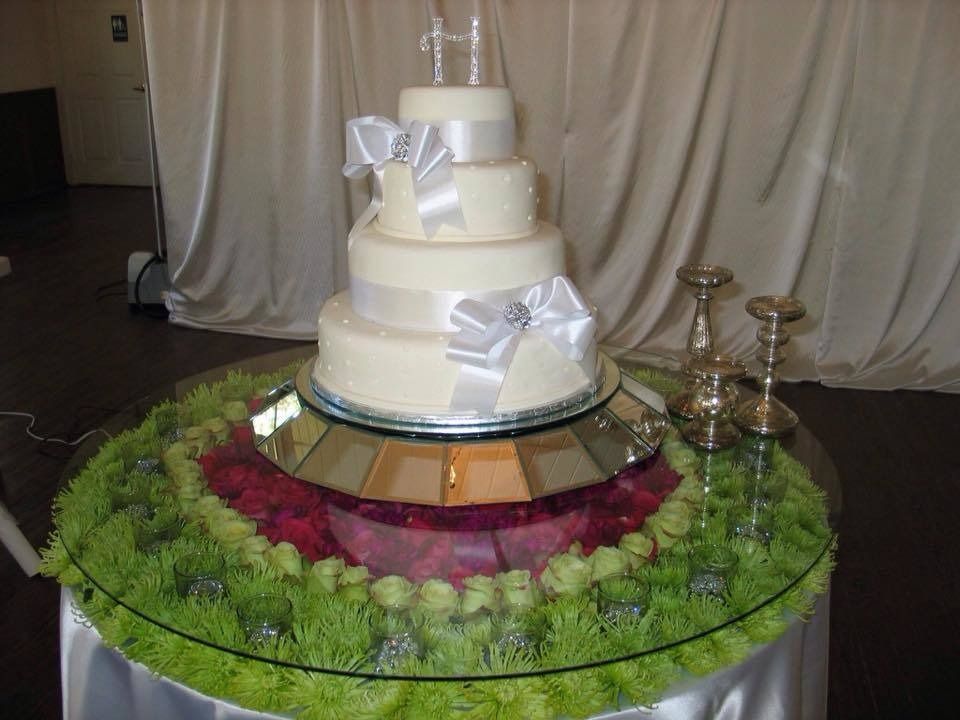 White tiered wedding cake on a decorated stand with a bow, surrounded by flowers and greenery.