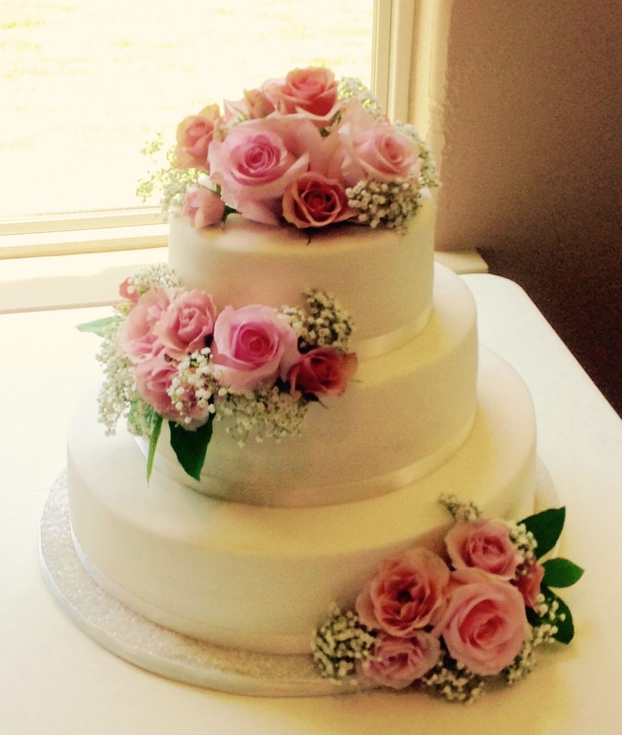 Three-tier white cake with pink rose decorations and white lace accents on a table