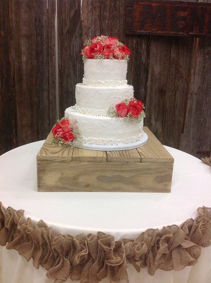 Three-tier white wedding cake with red flowers on a rustic wooden stand, on a white table with burlap trim