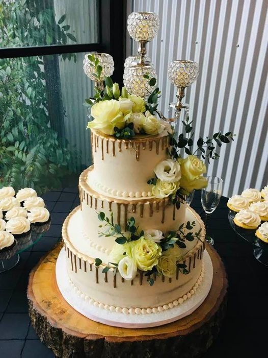 Three-tier white cake with gold drip, white and yellow flowers, on a round wooden stand