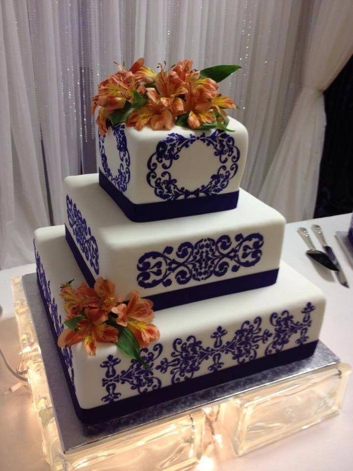 Three-tier white cake with purple scrollwork and orange flowers on a clear-lit stand