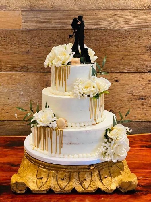 Three-tier white wedding cake with white flowers and gold drips on a carved wooden stand