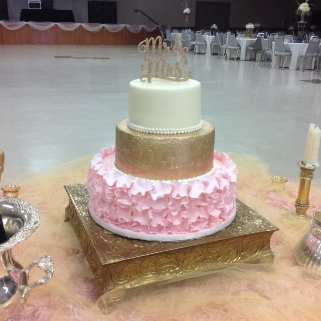 Three-tier white and gold wedding cake with pink ruffles on a gold stand in a banquet hall
