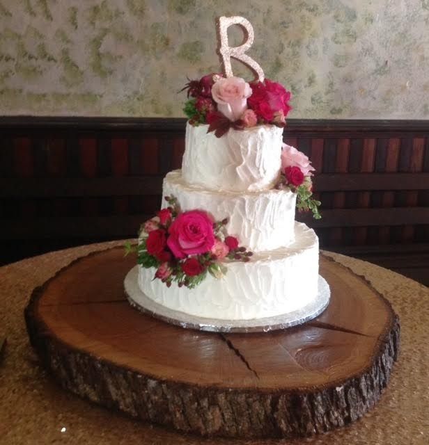 Three-tier white wedding cake with pink roses and a letter R topper on a wood slab cake stand
