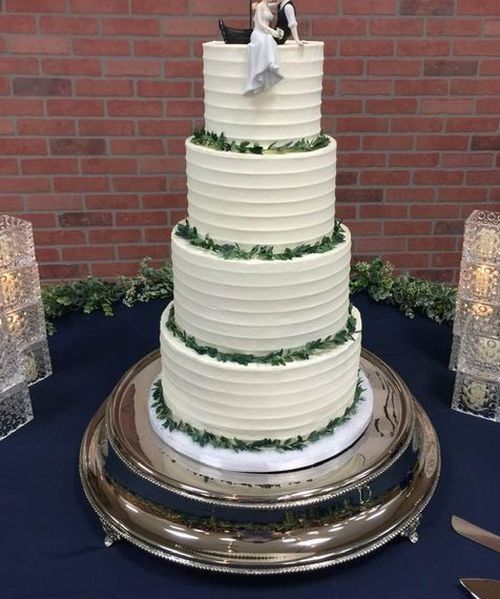 Four-tier white wedding cake with green garland accents on a silver stand against a brick wall.