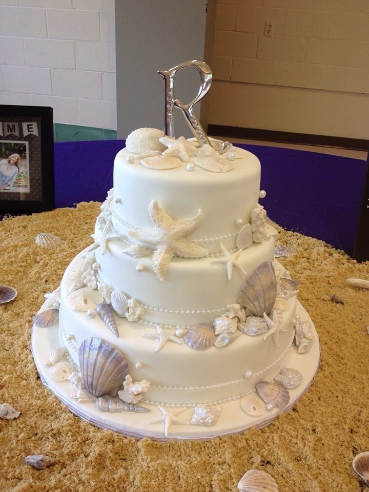 White tiered cake with seashell decorations and a silver letter R topper on a beach-themed table centerpiece