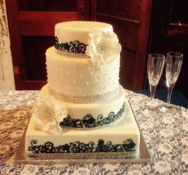 Three-tier white wedding cake with black lace decoration on a table, beside two champagne flutes