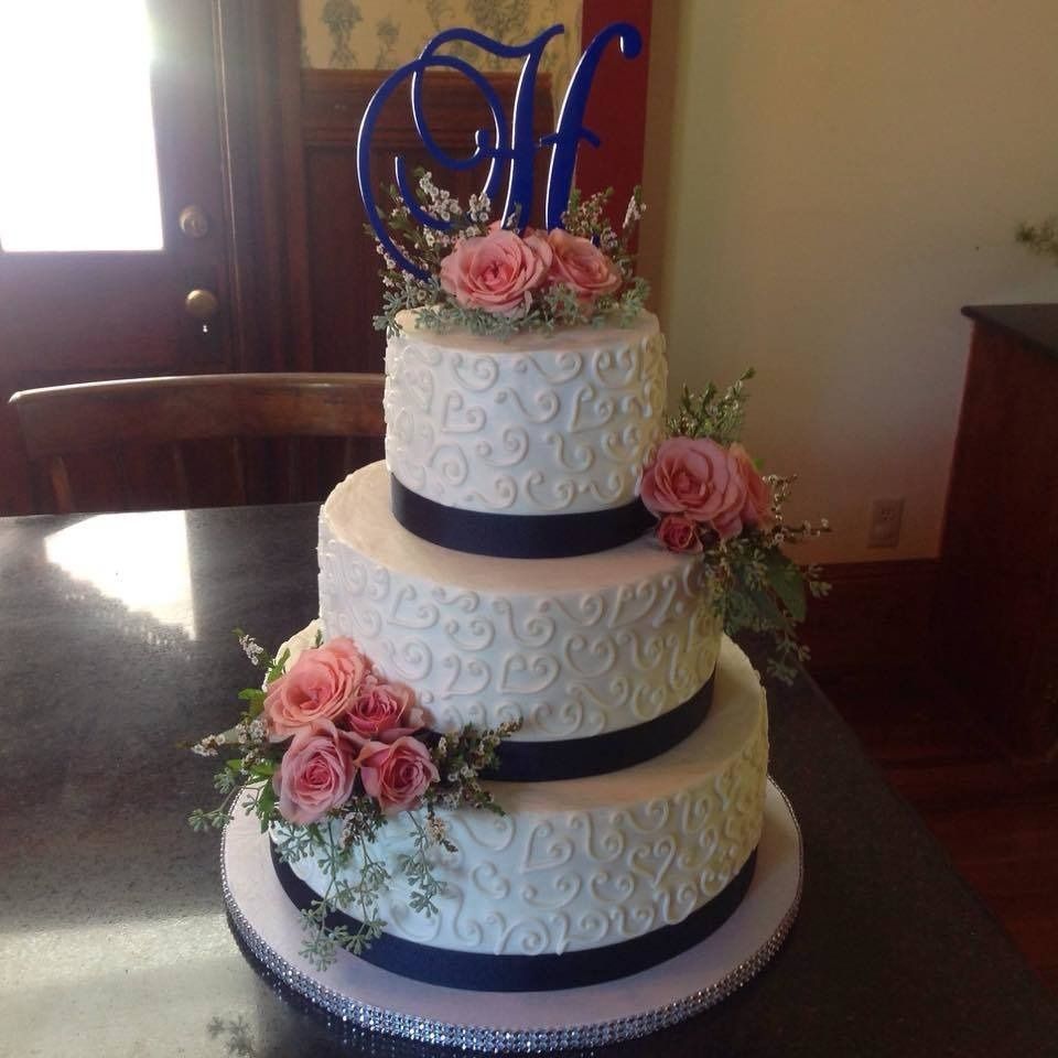 Three-tier white wedding cake with pink roses and a blue monogram topper on a table
