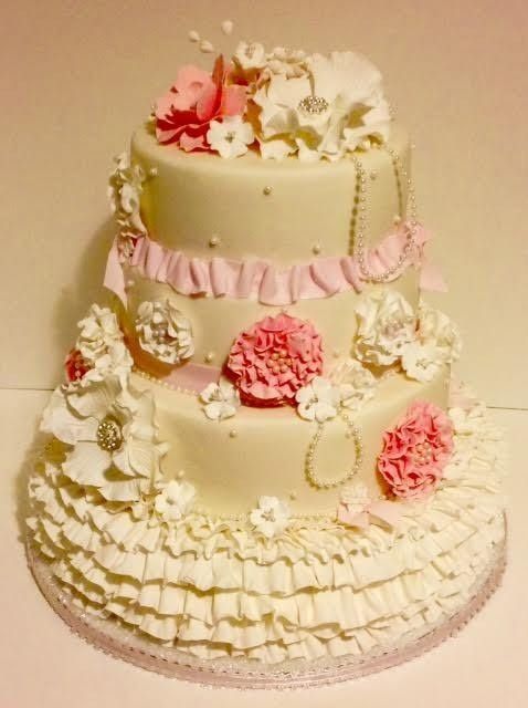 Two-tier white wedding cake with pink ribbon, ruffles, and floral decorations