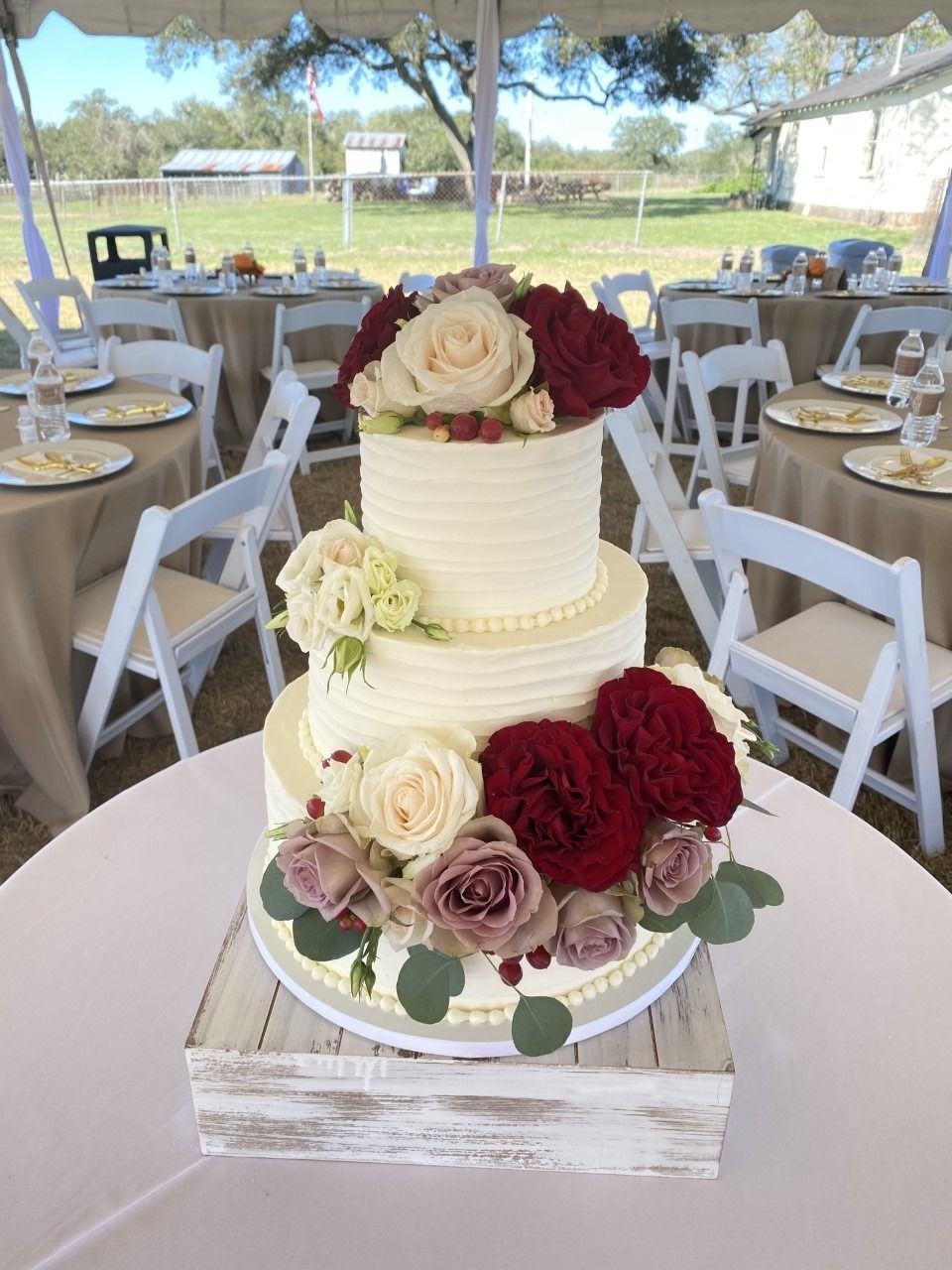 Three-tier white wedding cake with red and cream roses on a rustic stand at an outdoor reception