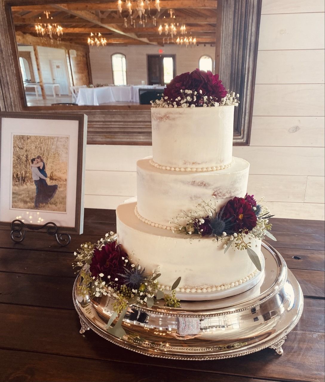 Three-tier white wedding cake with burgundy flowers on a silver tray beside a framed photo