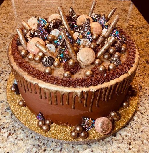 Chocolate drip cake topped with assorted candies, beads, and lollipops on a gold cake board