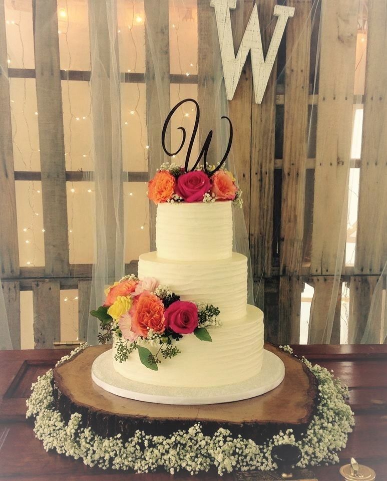 Two-tier white wedding cake with orange and pink flowers, topped with a black “W,” on a rustic wood backdrop.