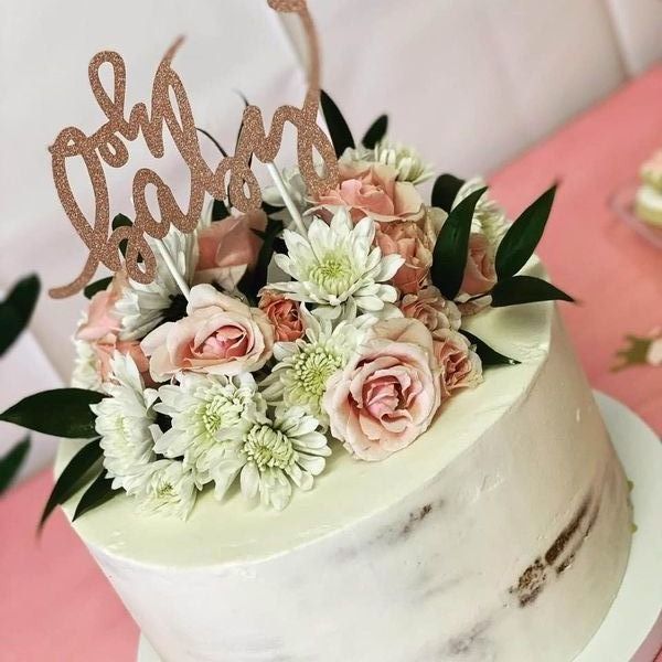 White cake with pink and white flowers and a gold “oh baby” topper on a pink surface