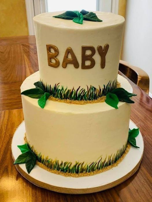 Two-tier white cake with gold “BABY” topper, green leaves, and grass-like trim on a wooden stand